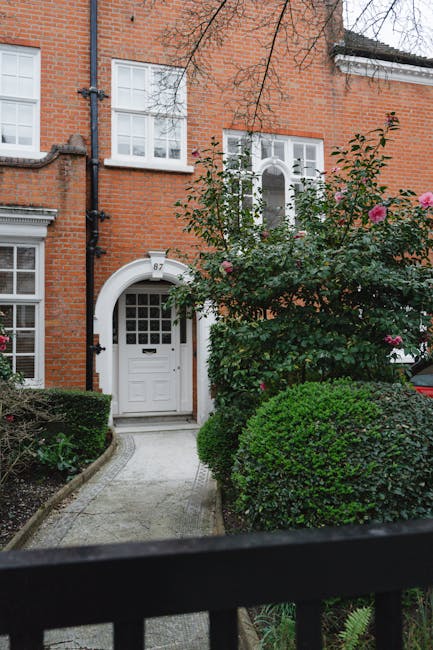 A residential garden scene featuring a narrow concrete pathway leading to a white front door within a brick house. The garden includes well-maintained, rounded green bushes and a flowering shrub with